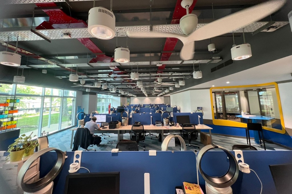 Interior of an office building with ceiling fans hanging above cubicles. Individual fans are visibile at each seat. One person is sitting in a cubicle midway through the room.