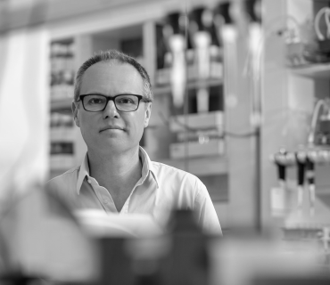 Gero Miesenböck, a white man with thin brown grey hair and glasses. He stands in a laboratory with out of focus pipettes in the foreground.