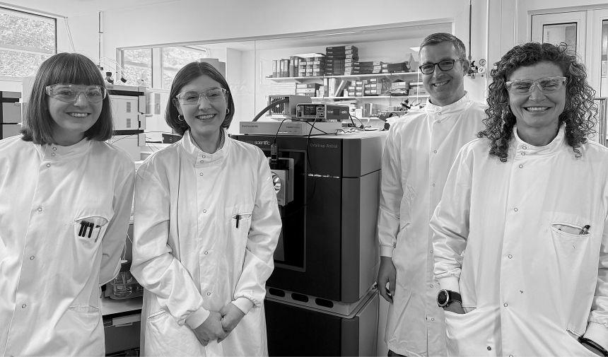 Three women and one man wearing white lab coats and clear safety glasses stand in a laboratory. Behind them is a table top analytical instrument contained in a large box-like structure.