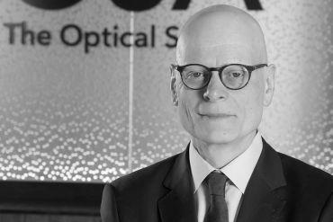 Portrait of Professor Ian Walmsley wearing glasses, dressed in a dark suit and patterned tie, against a blurred background featuring partial text reading 'The Optical'