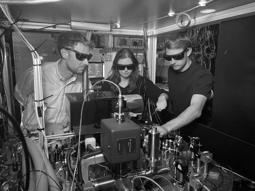 Two men and a young woman work on the interior of a quantum computer experimental set up. This has many instruments, cables and wires. They wear dark safety glasses.