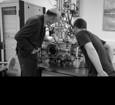 Two male scientists, with their backs to the camera, examine a complex scientific instrument on a lab bench.
