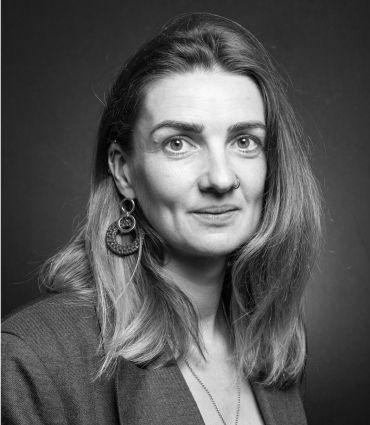 Professor Stephanie Hirmer, a white woman with shoulder-length brown hair. She stands in front of a spotlit plain dark background.