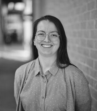 Dr Alycia Leonard, a white woman with long brown hair, wearing glasses. She stands in front of a brick wall.