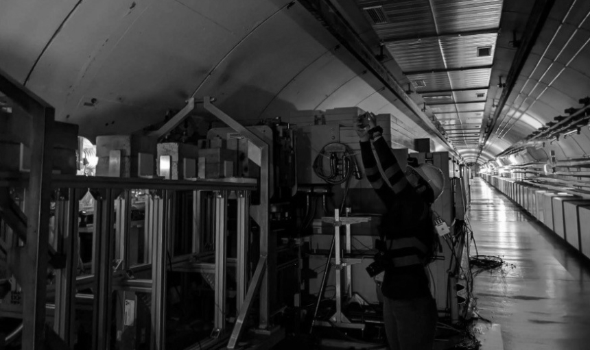 A researcher wearing a hard hat stands next to scientific machinery in a tunnel illuminated by orange and blue lighting.