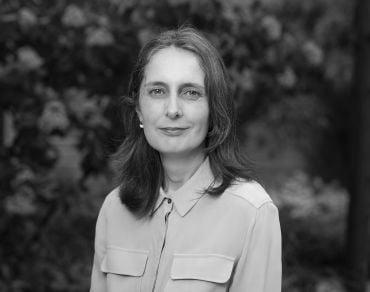 Portrait photograph of Rebecca Eynon, a white woman with long brown hair. She stands outside with a hedge in the background.
