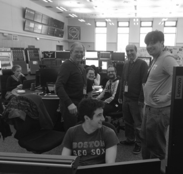 A group of male and female researchers sit and stand around a bank of computers in the CERN control room.