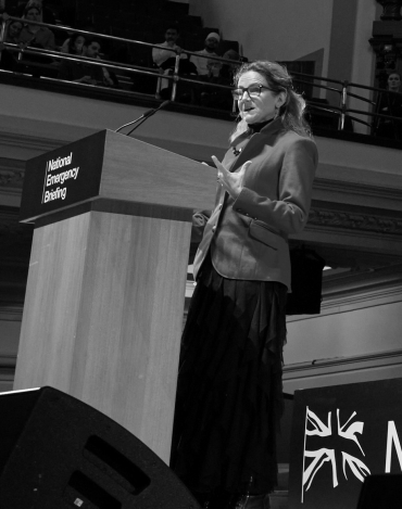 A white woman with blonde hair wearing a red jacket and black skirt stands at a lectern branded 'National Emergency Briefing'