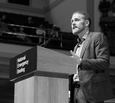 A white man wearing a grey-blue suit speaks at a lectern branded with 'National Emergency Briefing.'