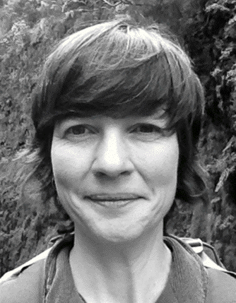 Black and white portrait photo of a woman with shoulder length hair. She stands outside in front of a hedge.
