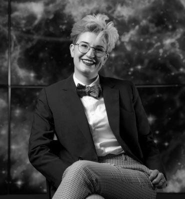 Portrait of Dr Héloïse Stevance, a young lady with curly orange and red hair, wearing a dark blue jacket, glasses and a colourful bow tie. She sits in front of a colourful outer-space backdrop.