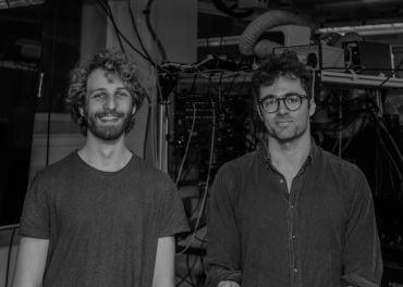 Two white men wearing blue shirts stand in a laboratory in front of apparatus for quantum computing.