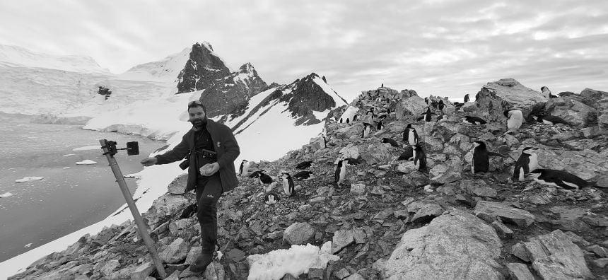 A man wearing a warm red coat stands next to a camera mounted on a tripod on a rocky shoreline, with icy mountains and the Antarctic sea in the background. Around him are several black and white penguins.
