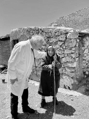 A man wearing a white coat stands with an elderly lady wearing a hooded coat and holding onto a staff, outside a stone dwelling.