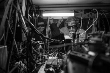 Two male researchers wearing safety spectacles look into the interior of a quantum computing experiment seen as many wires and connections.