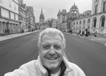 Peter Grindrod, a white man with white hair, standing in Oxford High Street with The Queen’s College and the University Church of St Marys visible.