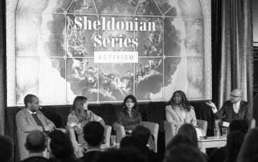 Moderator Dr Julius Grower addresses the panellists on stage with the Sheldonian Series graphic on screen behind them