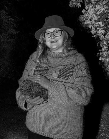 A lady with long blonde hair wearing a green jumper and a brown hat holds a hedgehog in an outdoor area at night.