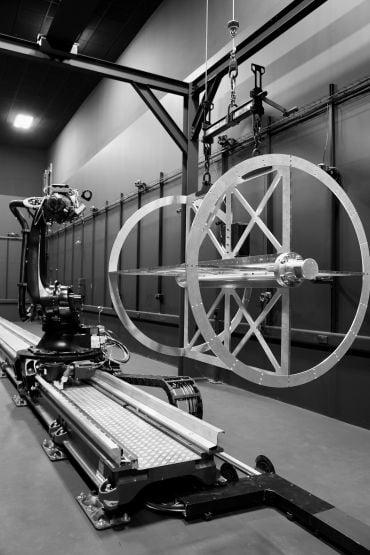 A circular metal frame (component of a satellite) is suspended from a metal rack. Behind it is a robotic arm used in manufacturing.