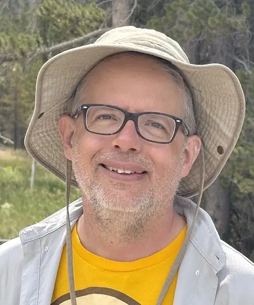 A man wearing a tan bucket hat, glasses and a white button-up shirt over a yellow T-shirt smiles at the camera.