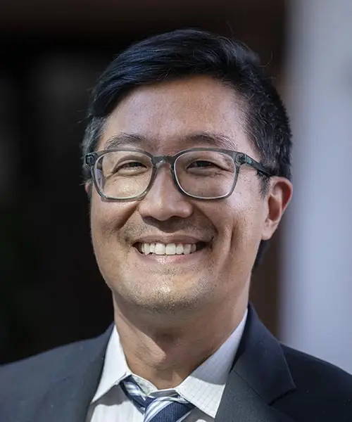 A man wearing glasses and a suit with a blue tie, white collared shirt and gray jacket smiles for the camera.