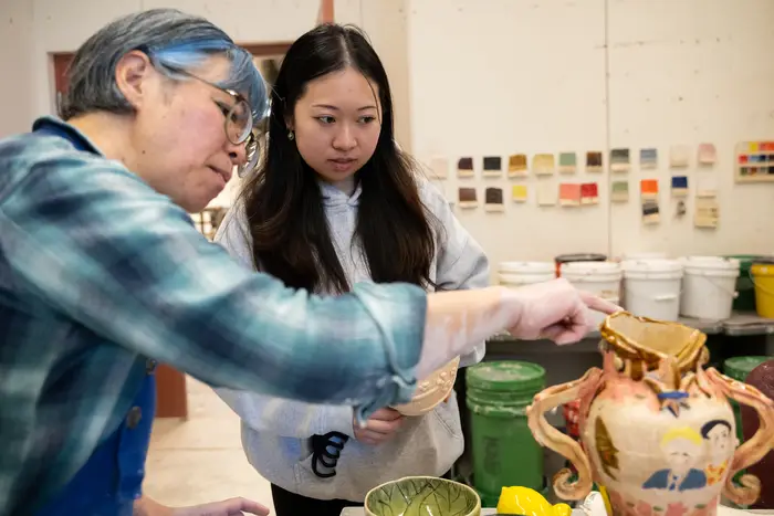 A woman wearing a blue plaid shirt and a blue apron gestures toward a ceramic pot as a student looks on intently.