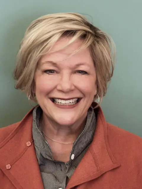 Headshot of a smiling woman with ear-length blonde hair wearing a burnt orange jacket and green scarf in front of a teal wall.