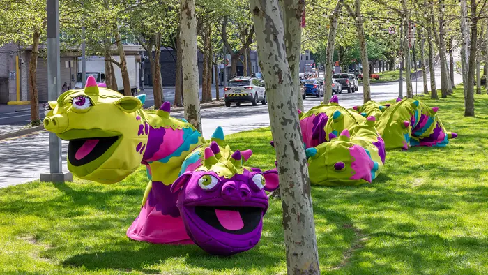 A sculpture resembling a large green and purple worm-like friendly monster sits in sections among trees in a long grassy median space alongside a road.