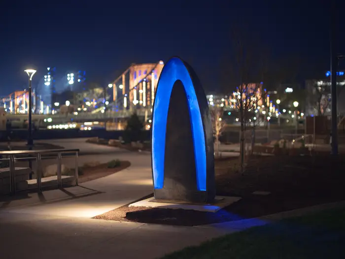 An blue inverted U-shape is illuminated and in focus in the foreground of a nighttime photo with one of Pittsburgh's bridges lit by streelights in the background.