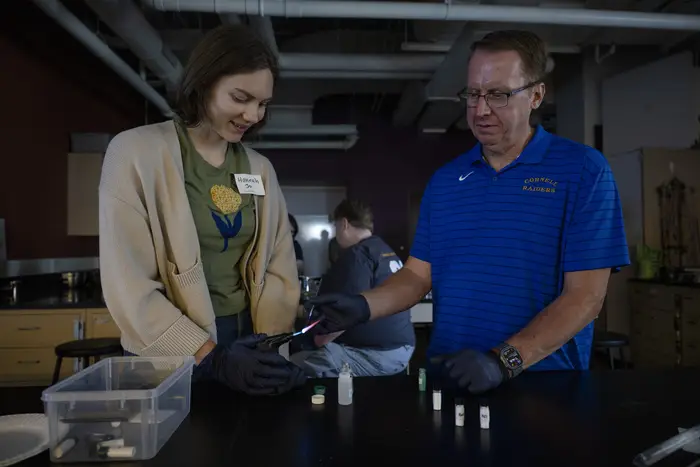 A woman in a brown cardigan and green T-shirt with short brown hair and a man in a blue polo shirt with short brown hair and glasses work together in a lab. The woman holds a butan lighter and the man holds a lab instrument to burn salts in the flame.